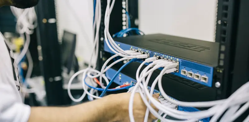 Technician patching white ethernet cables into a network switch in a comms rack