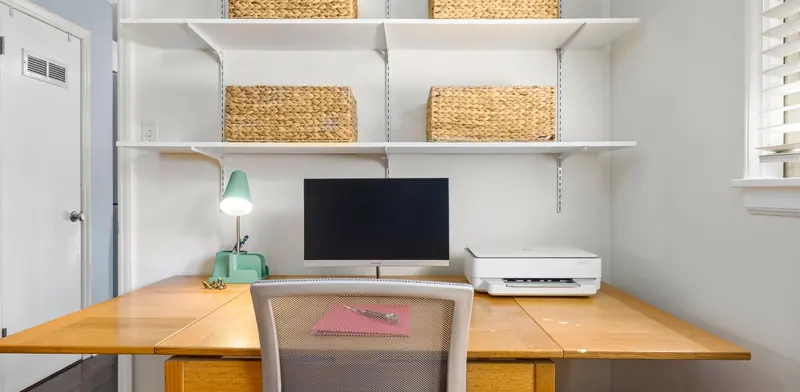 Home office desk with a desktop computer, printer and shelving in a bright room